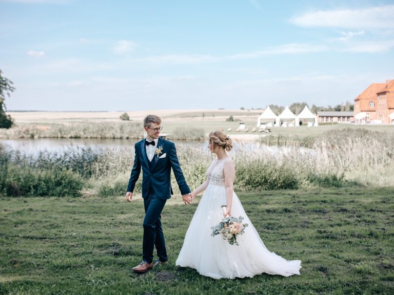 Brautpaar hält Händchen und läuft an einem Gewässer entlang, umgeben von Natur.